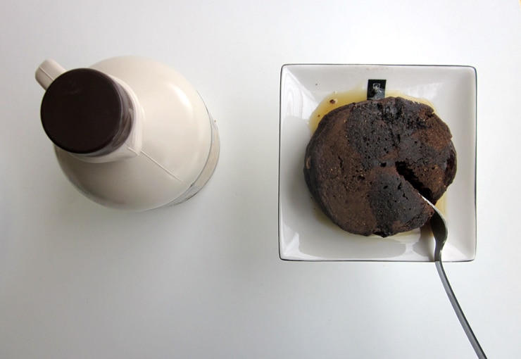 A fork cutting into chocolate pancakes on a white plate next to a container of maple syrup.