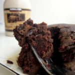 A fork holding up a bite of chocolate pancakes on a white plate next to a container of maple syrup.