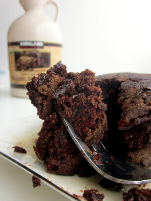 A fork holding up a bite of chocolate pancakes on a white plate next to a container of maple syrup.