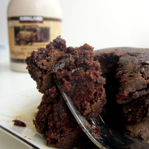 A fork holding up a bite of chocolate pancakes on a white plate next to a container of maple syrup.
