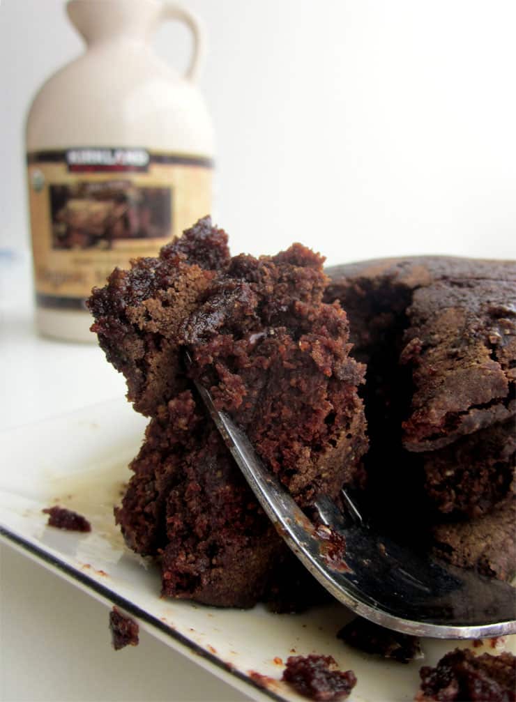 A fork holding up a bite of chocolate pancakes on a white plate next to a container of maple syrup.
