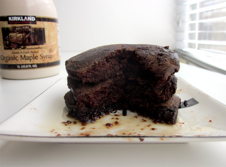 A wedge taken out of a stack of 3 chocolate pancakes on a white plate next to a container of maple syrup.