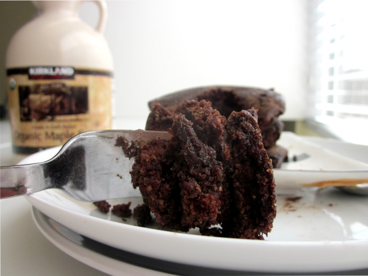 A fork holding a bite of chocolate pancakes on a white plate next to a container of maple syrup.