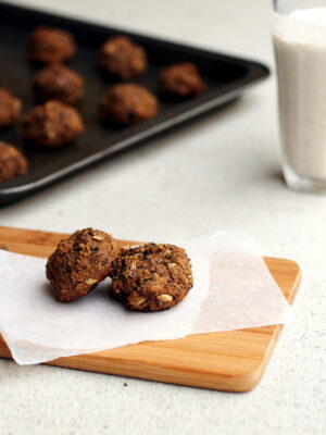 Two grainy cookies on a piece of parchment paper on a cutting board with a baking tray full of cookies in the background.