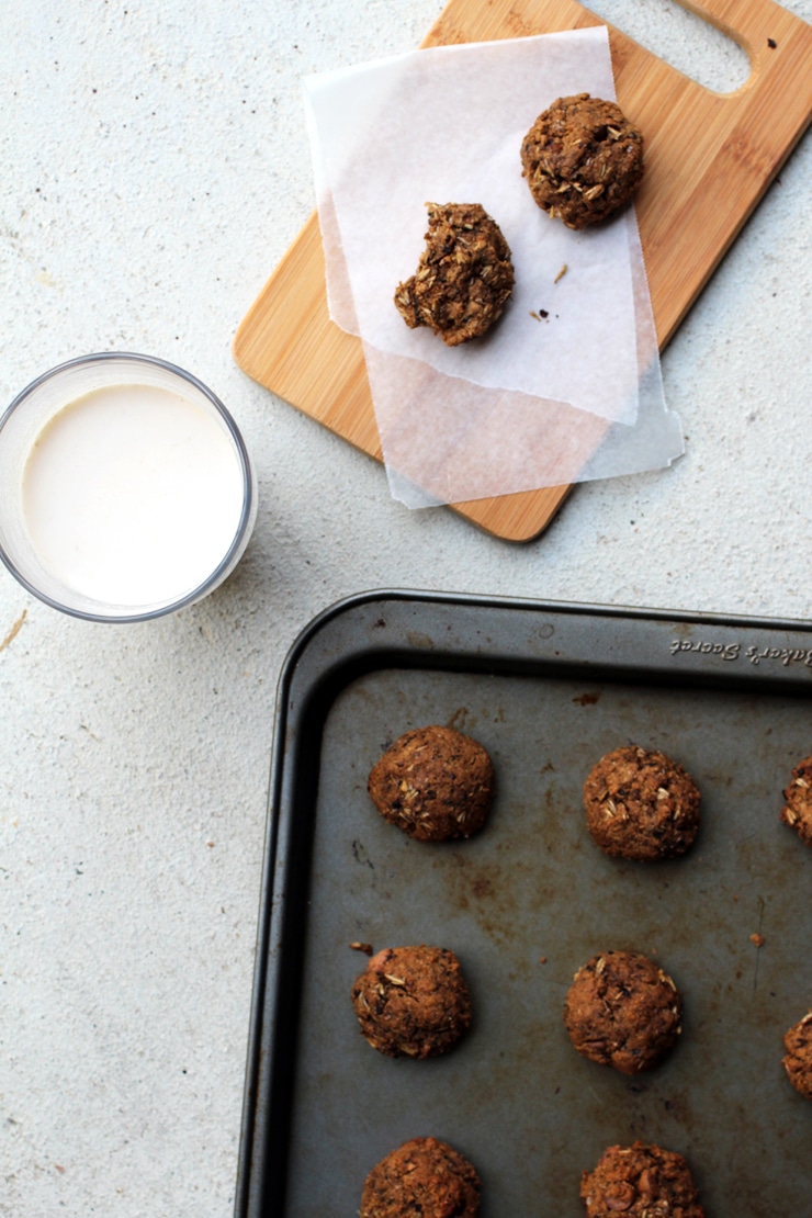 A half-eaten cookie on a piece of parchment paper on a cutting board next to a baking tray full of cookies and a glass of milk.