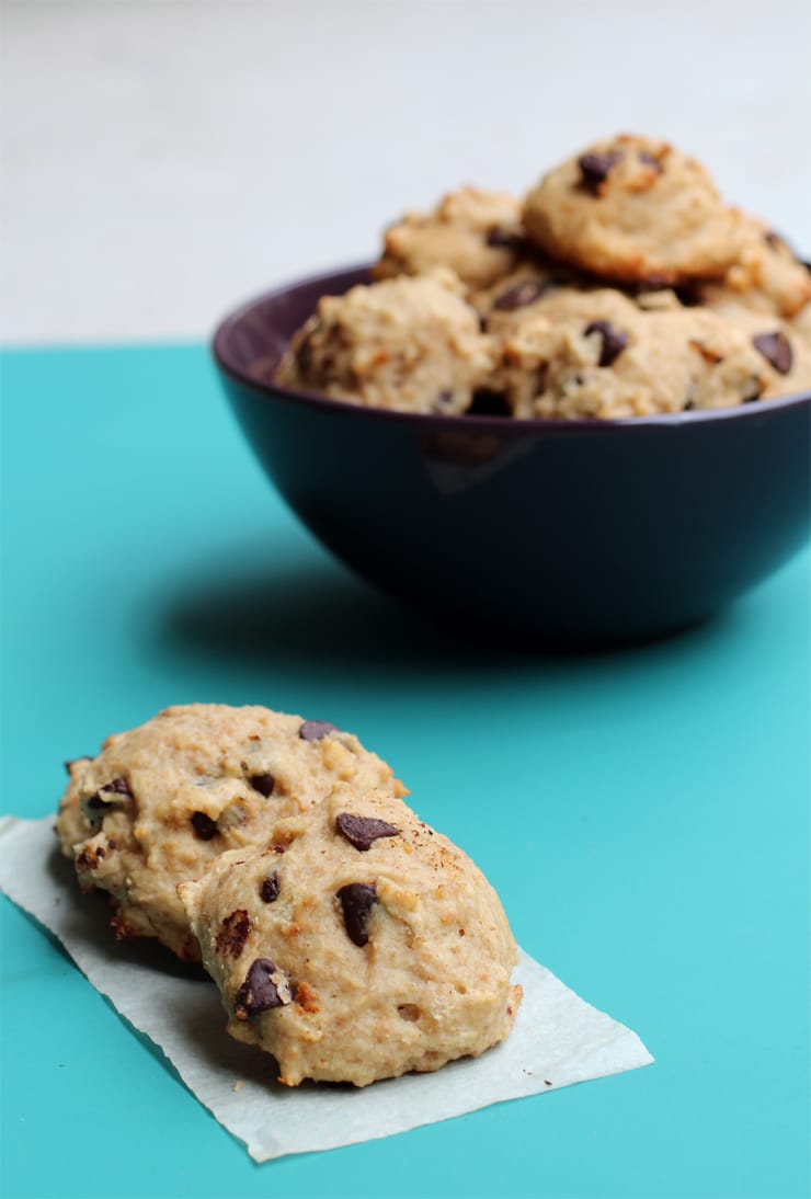 A purple bowl full of banana chocolate chip cookies with two cookies on a piece of parchment paper in the foreground.