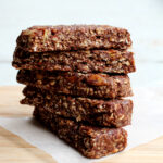 Five date-sweetened granola bars stacked on a sheet of parchment paper on a cutting board.