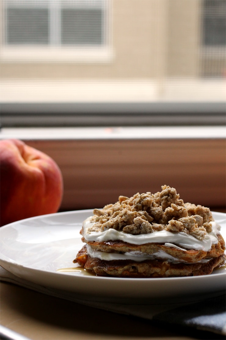 A stack of pancakes on a white plate layered with yogurt, peach slices and granola.