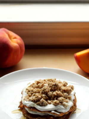 A stack of pancakes on a white plate layered with yogurt, peach slices and granola with peaches in the background.