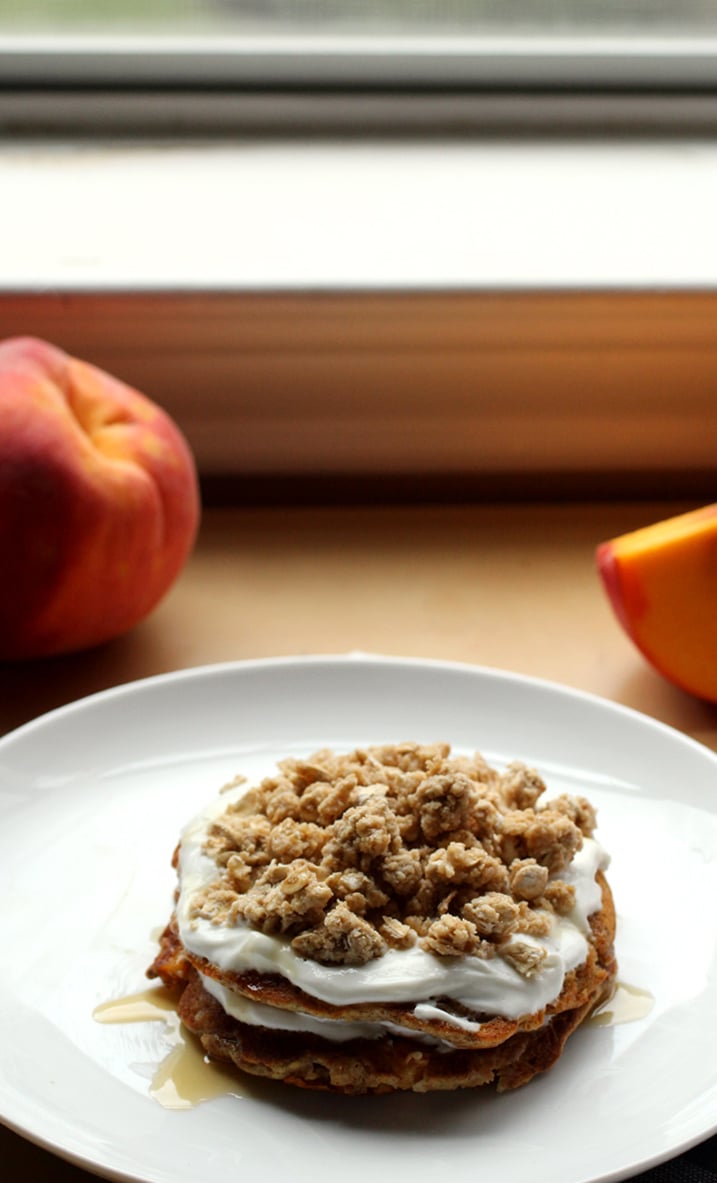 A stack of pancakes on a white plate layered with yogurt, peach slices and granola with peaches in the background.
