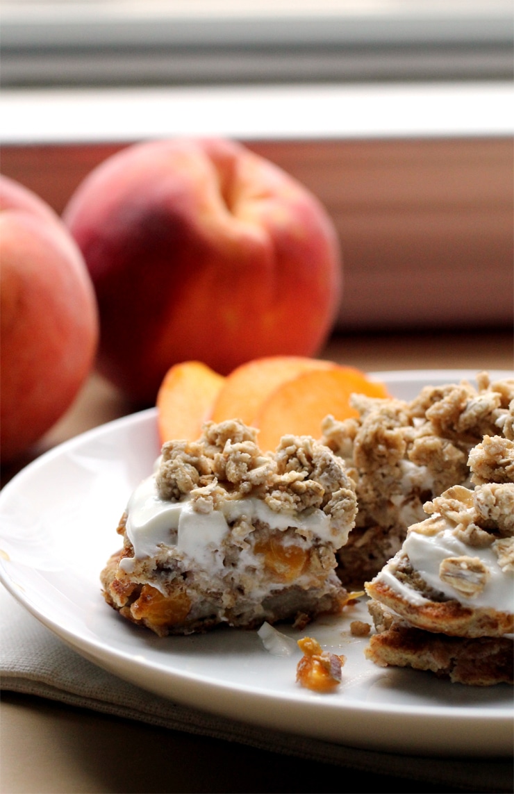 A stack of pancakes on a white plate layered with yogurt, peach slices and granola with a bite taken out of it.