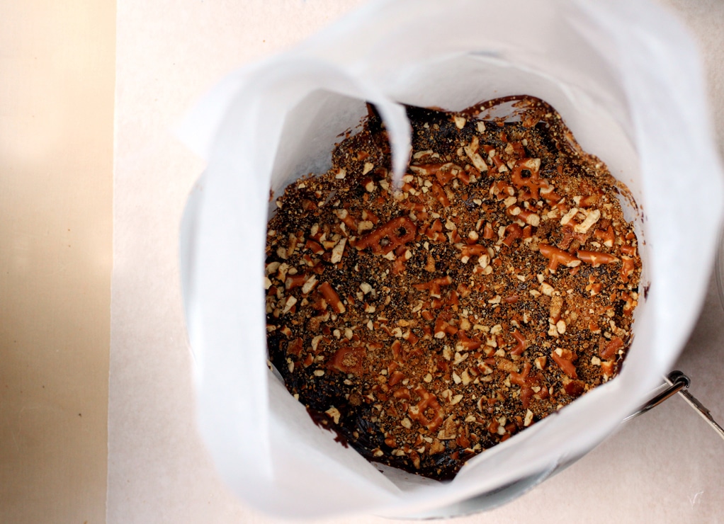 A cake pan lined with parchment paper and filled with a layer of cake, chocolate ganache and pretzel crumbles.