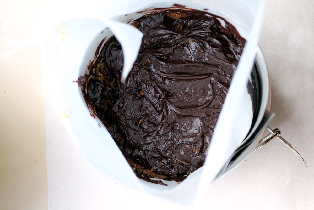 A cake pan lined with parchment paper and filled with a layer of cake and chocolate ganache.