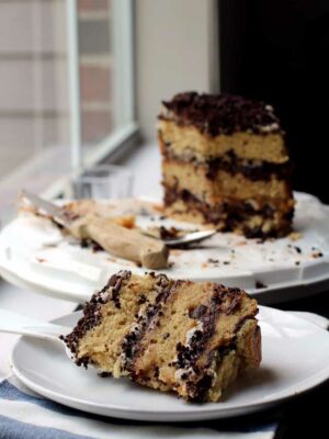 A slice of a triple layer chocolate chip cake layered with chocolate crumbs on a white plate with the rest of the cake in the background.