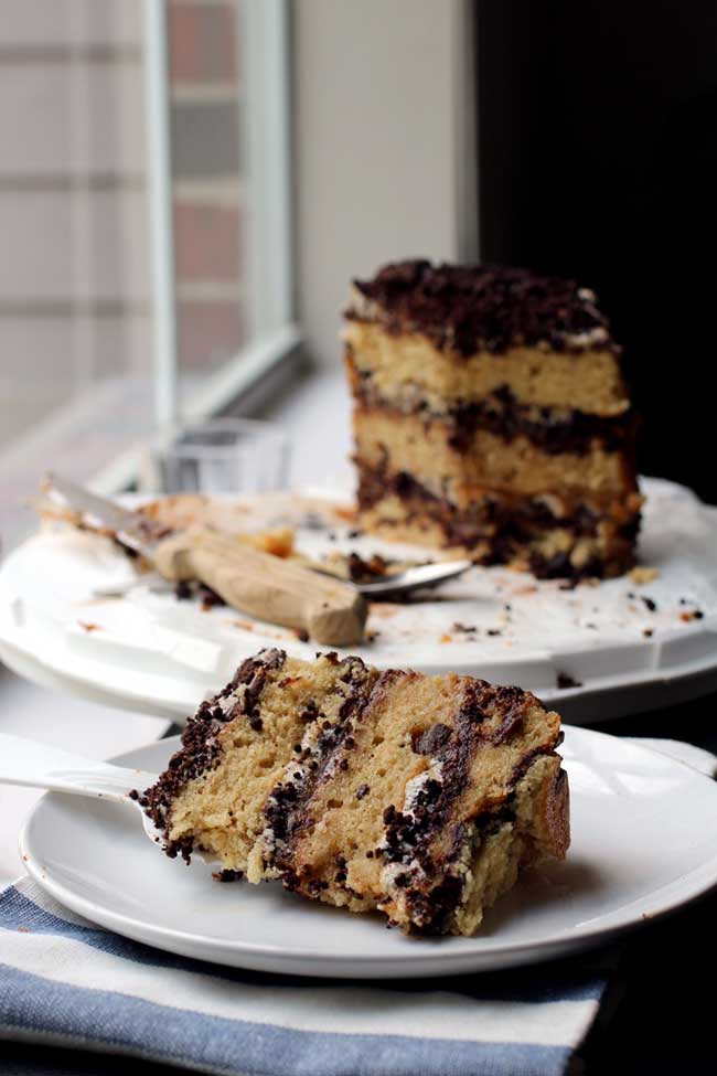 A slice of a triple layer chocolate chip cake layered with chocolate crumbs on a white plate with the rest of the cake in the background.