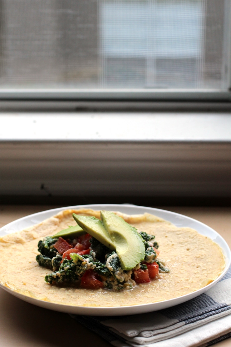 A chickpea flour tortilla on a white plate topped with kale, avocado and carrots.