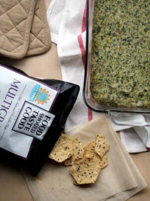 A glass pan of spinach artichoke dip next to a bag of crackers spilling out of the bag onto a towel.