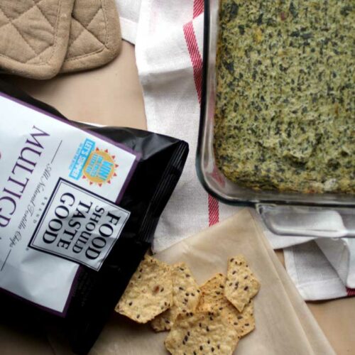 A glass pan of spinach artichoke dip next to a bag of crackers spilling out of the bag onto a towel.
