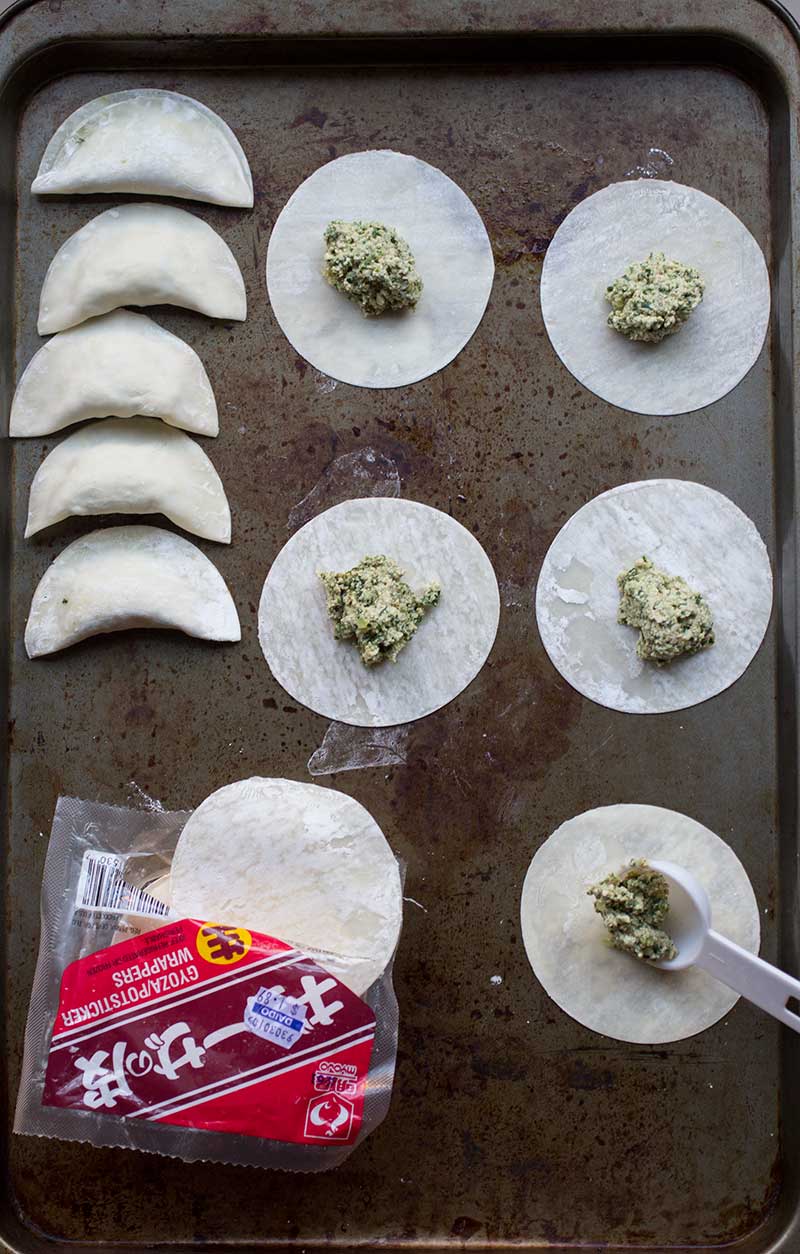 Assembled tofu kale dumplings on a metal tray next to rounds of wrappers filled with balls of filling.