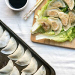 Tofu kale potstickers on top of a large piece of lettuce on a cutting board next to a bowl of soy sauce.