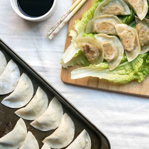 Tofu kale potstickers on top of a large piece of lettuce on a cutting board next to a bowl of soy sauce.