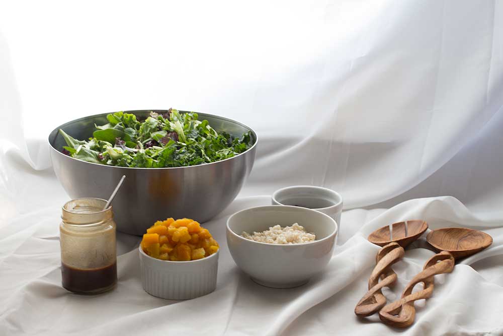 A silver bowl filled with mixed greens next to ramekins filled with butternut squash, farro, salad dressing and wooden tongs.