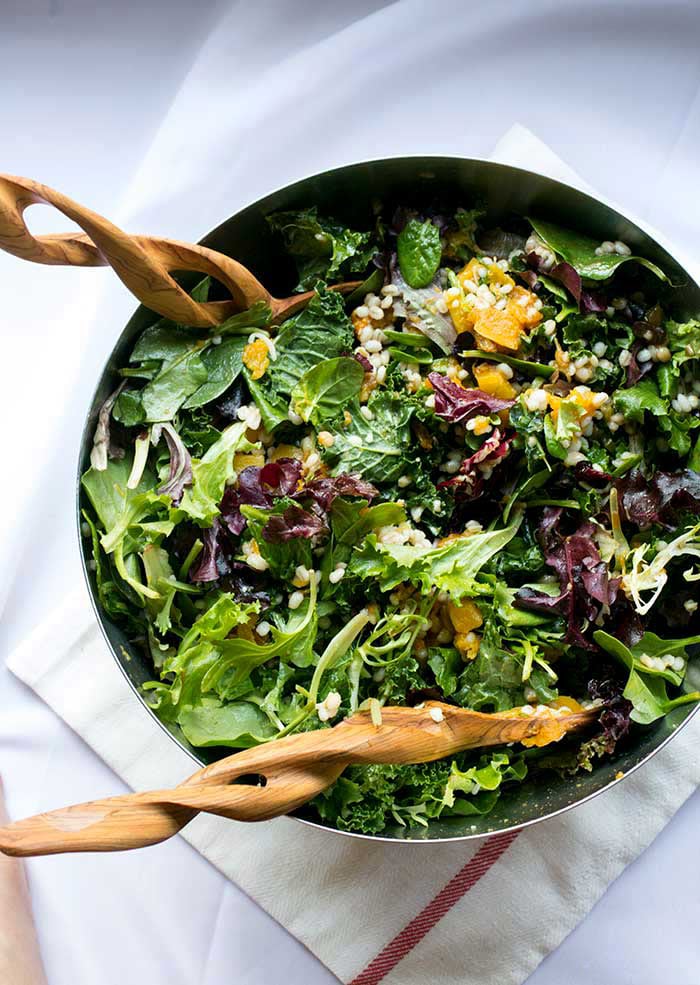 A silver bowl full of mixed greens with two wooden salad tongs.