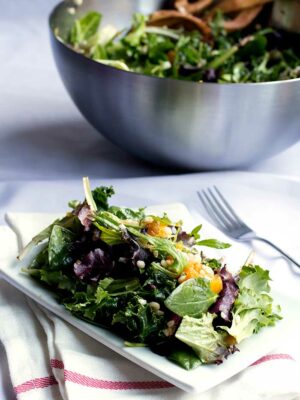 A white plate filled with mixed greens with a silver bowl full of salad in the background.