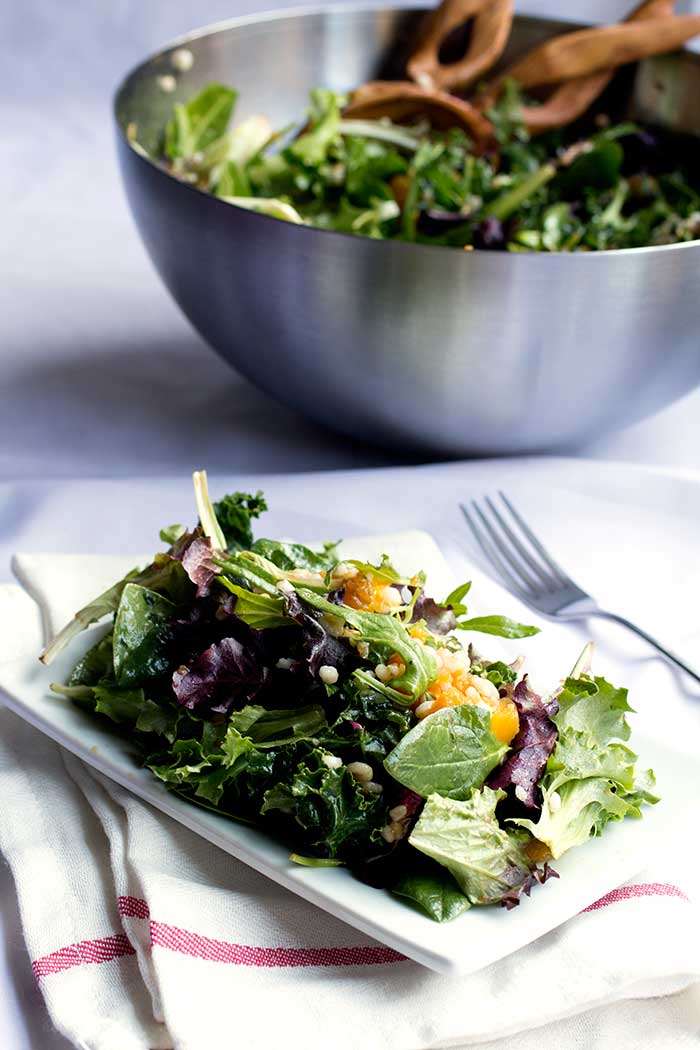 A white plate filled with mixed greens with a silver bowl full of salad in the background.