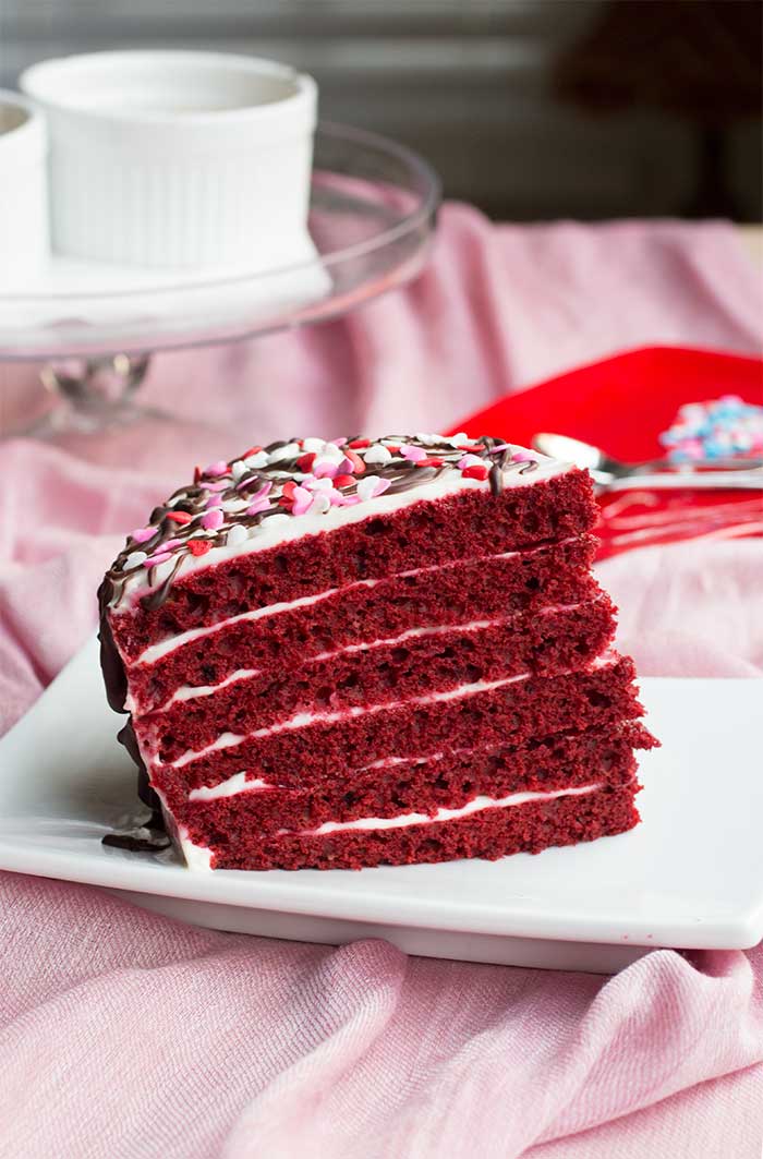 A six layer slice of red velvet cake topped with sprinkles and chocolate drizzles.
