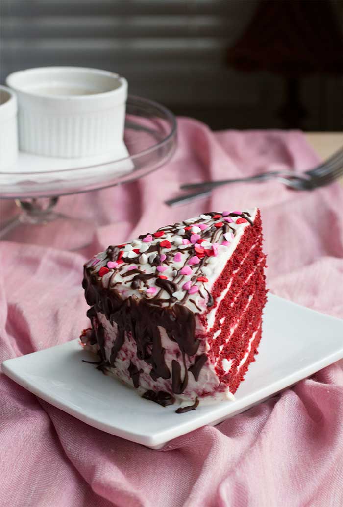 A slice of 6-layer red velvet cake with sprinkles and chocolate drizzles on a white plate on top of a pink tablecloth.
