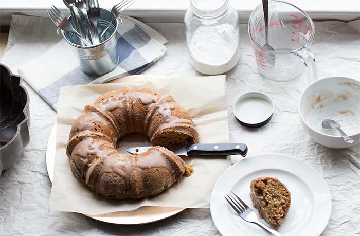 A frosted bundt cake on a piece of parchment next to a slice of cake on a white plate with a fork.