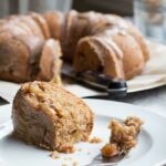 A slice of apple bundt cake on a white plate with a bundt cake in the background.