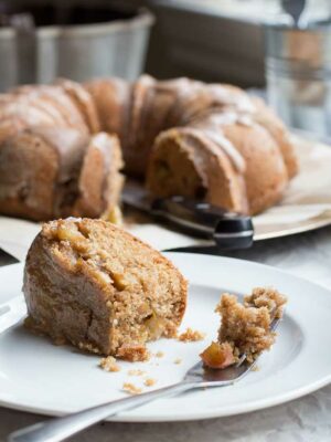 A slice of apple bundt cake on a white plate with a bundt cake in the background.