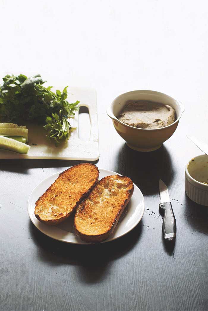 Two halves of a toasted bread roll on a white plate next to a knife, a bowl of lentil pate and herbs on a cutting board.