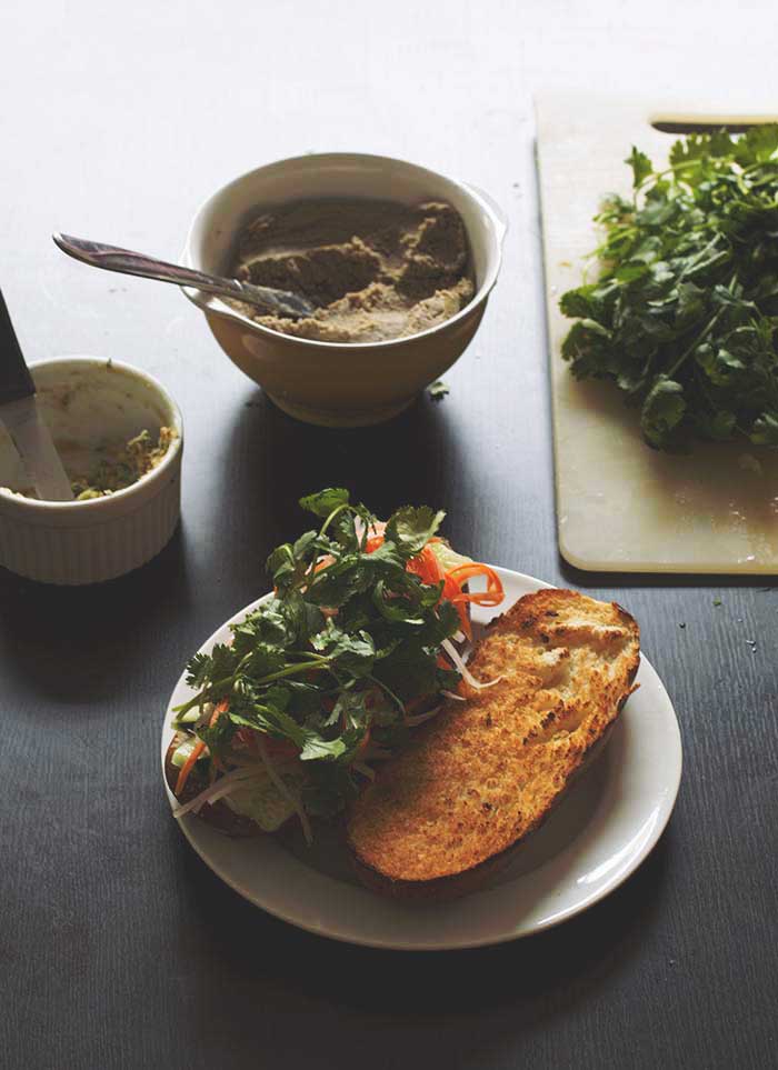 A banh mi on a white plate with the top half of bread on the plate with a bowl of lentil pate in the back.