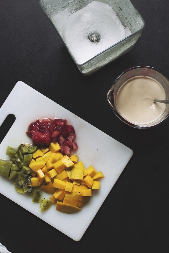 Chopped kiwi, mango and strawberries on a cutting board next to a cup of condensed milk.