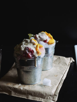 Two silver mini pails filled with shaved ice, chopped mango and strawberries and topped with condensed milk.