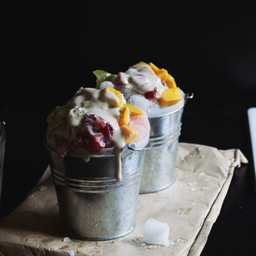 Two silver mini pails filled with shaved ice, chopped mango and strawberries and topped with condensed milk.