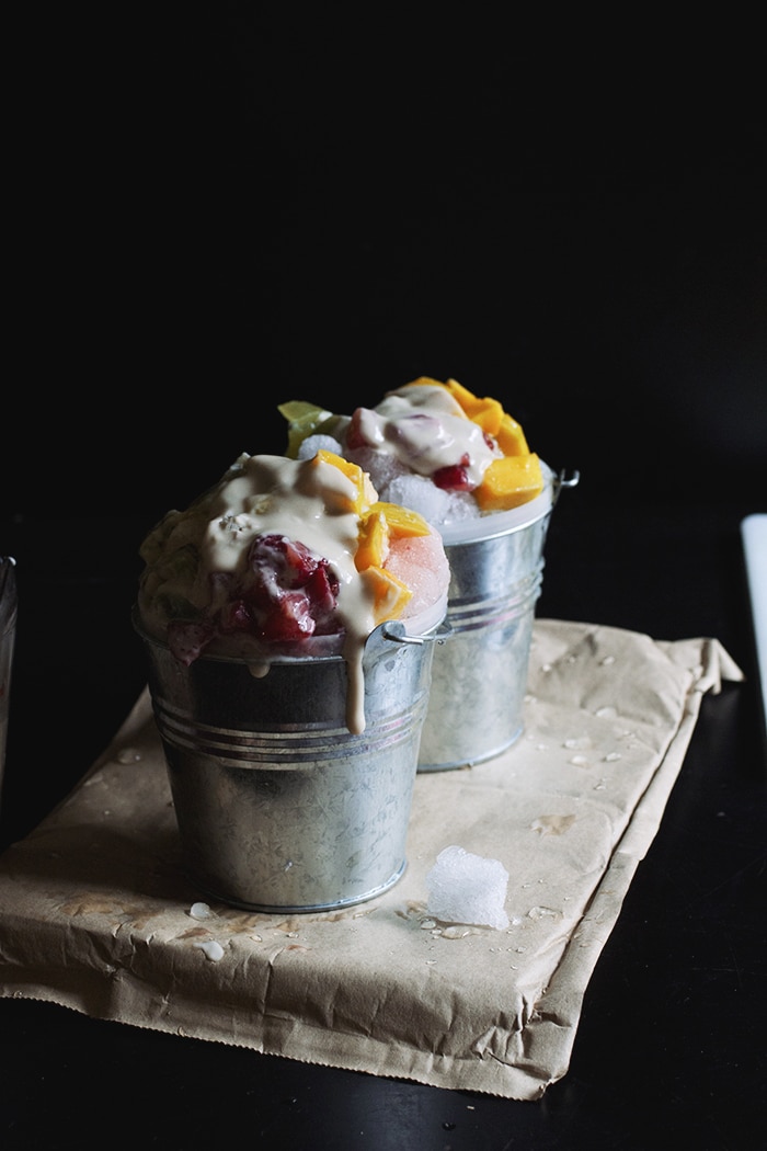 Two silver mini pails filled with shaved ice, chopped mango and strawberries and topped with condensed milk.