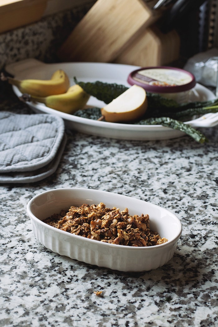 granola in a white bowl on a marble countertop with fruit in the background.