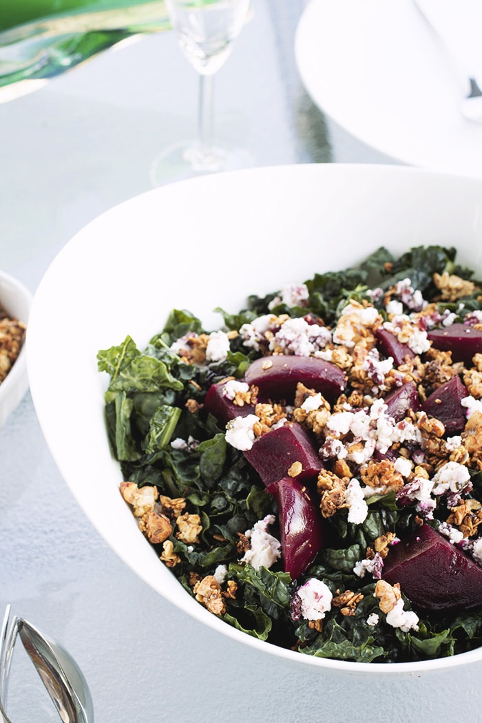 white bowl filled with kale, goat cheese, beets and walnut granola on a glass table.
