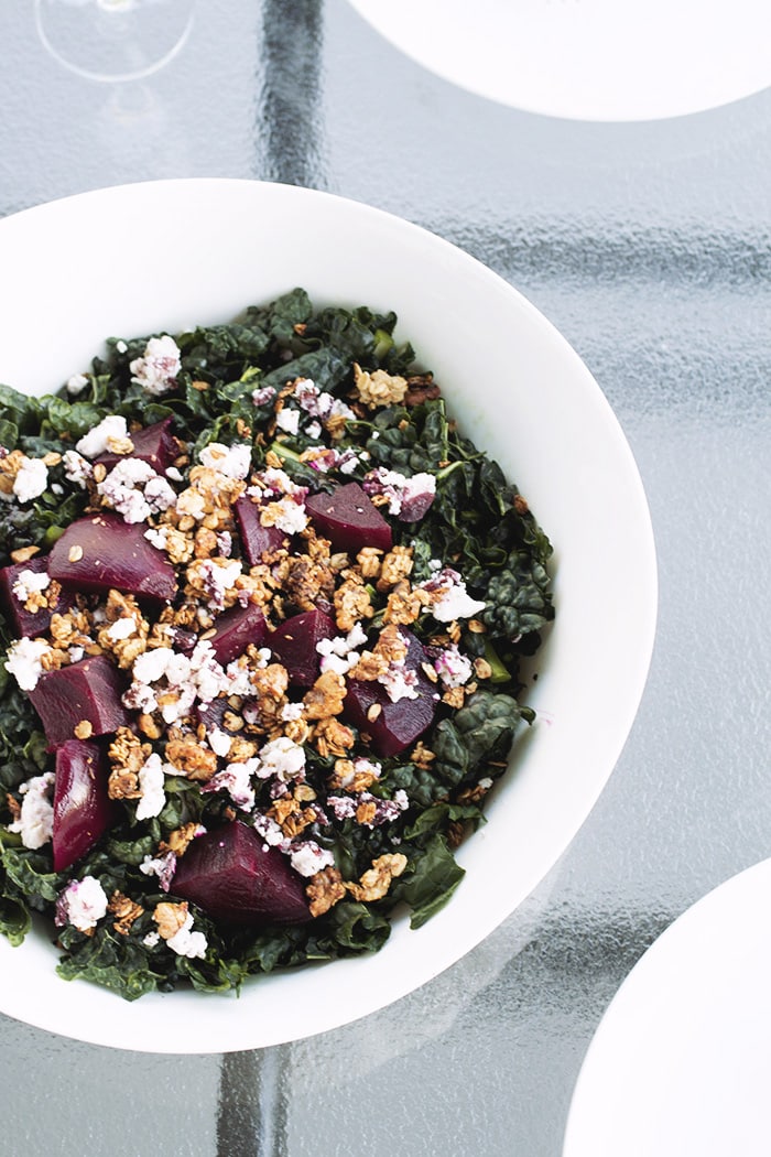 White bowl filled with kale, goat cheese, beets and walnut granola on a glass table.