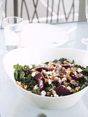 white bowl filled with kale, goat cheese, beets and walnut granola on a glass table.