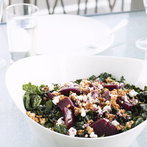 white bowl filled with kale, goat cheese, beets and walnut granola on a glass table.
