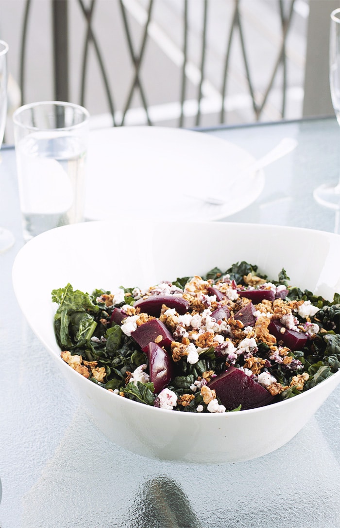 white bowl filled with kale, goat cheese, beets and walnut granola on a glass table.
