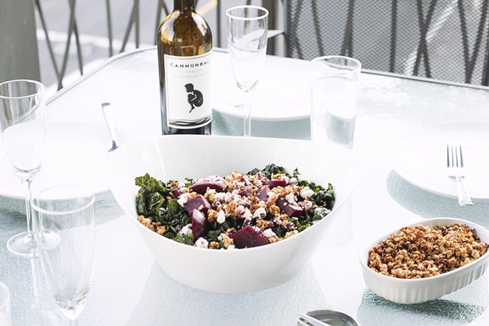white bowl filled with kale, goat cheese, beets and walnut granola on a glass table with wine glasses and other plates.