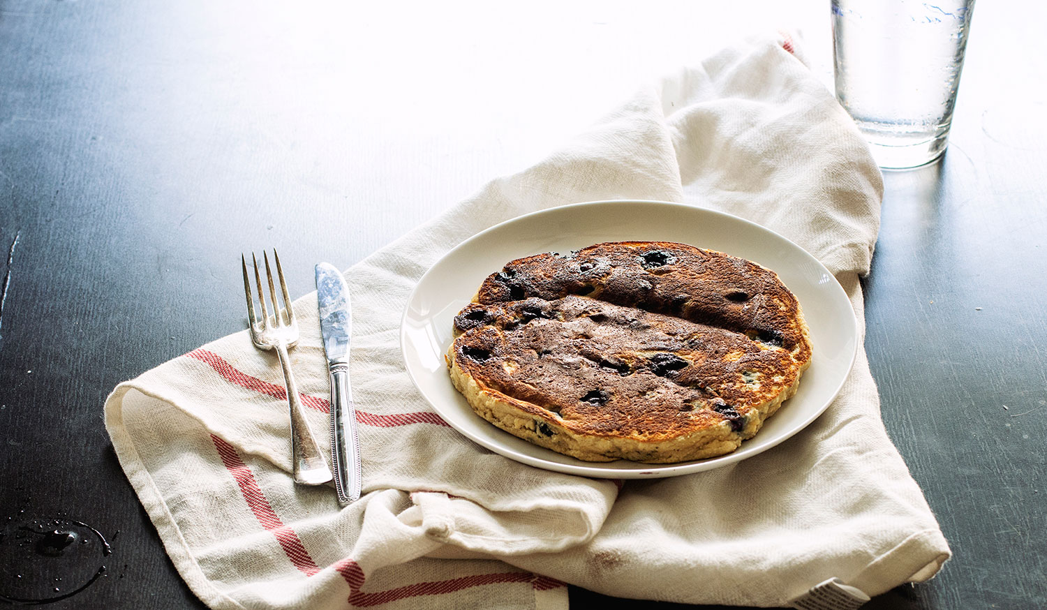 A thick blueberry pancake on a white plate on top of a white towel with a fork and knife on the table.