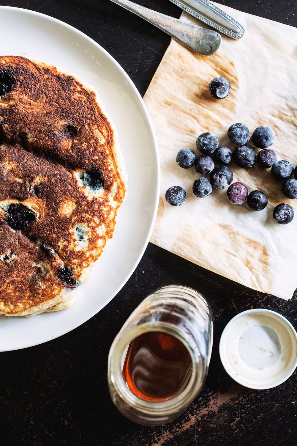 A thick blueberry pancake on a white plate with a slice taken out of it next to blueberries on a black table.