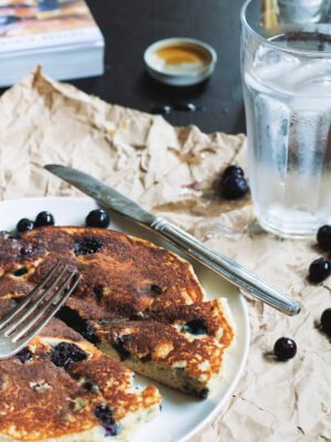 A thick blueberry pancake on a white plate with a slice taken out of it.